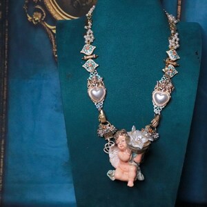 May include: Ornate necklace featuring a cherub pendant holding flowers. The necklace has heart-shaped pendants with pearl accents, and decorative elements in gold, teal, and pink. The necklace is displayed on a green mannequin.