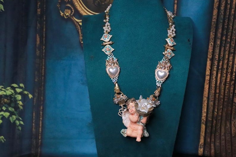 May include: Ornate necklace featuring a cherub pendant holding flowers. The necklace has heart-shaped pendants with pearl accents, and decorative elements in gold, teal, and pink. The necklace is displayed on a green mannequin.