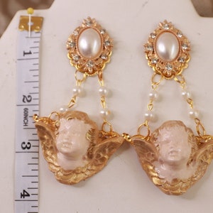 以下が含まれることがあります： ゴールドトーンのイヤリング。ケルビムのチャーム、パールアクセント、ゴールドセッティングの大きなフェイクパールが特徴です。