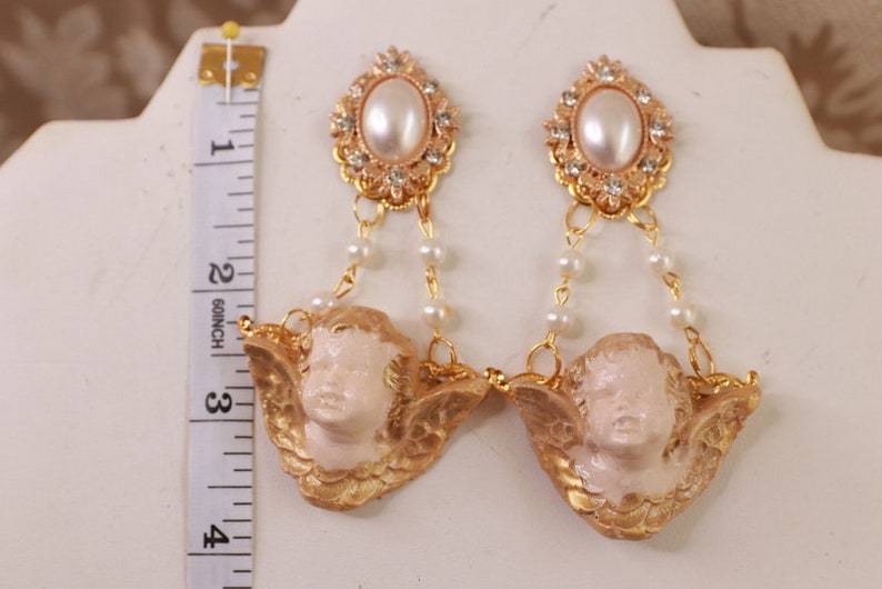 以下が含まれることがあります： ゴールドトーンのイヤリング。ケルビムのチャーム、パールアクセント、ゴールドセッティングの大きなフェイクパールが特徴です。