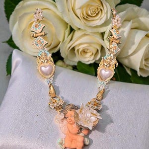 May include: A gold-toned necklace featuring a cherub holding a flower, with heart-shaped pendants and ornate detailing. The necklace is adorned with pearls and turquoise accents, set against a backdrop of white roses.