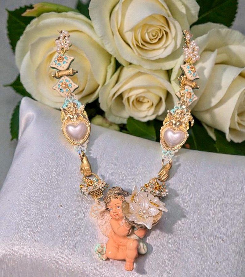 May include: A gold-toned necklace featuring a cherub holding a flower, with heart-shaped pendants and ornate detailing. The necklace is adorned with pearls and turquoise accents, set against a backdrop of white roses.