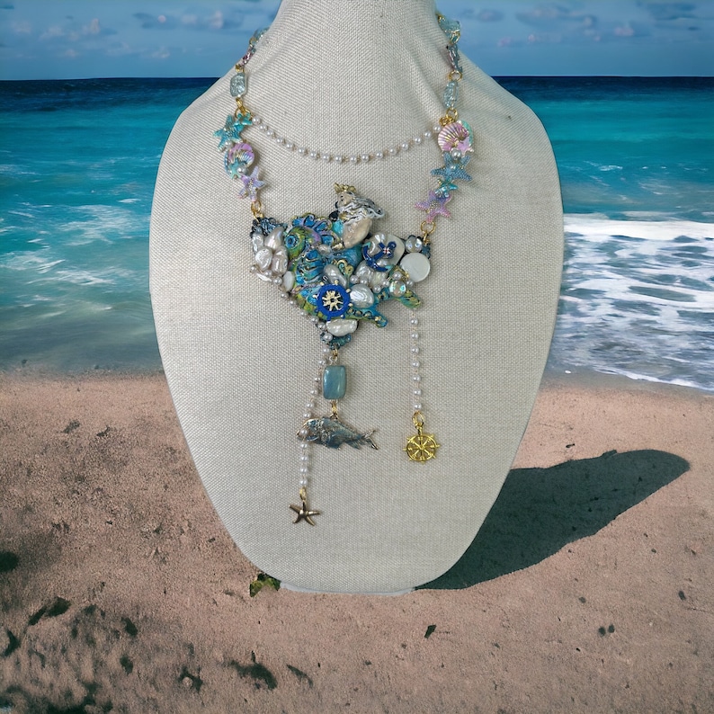 May include: A blue and white seashell necklace with a pearl and gold chain. The necklace features a variety of seashells, starfish, and a small fish charm.