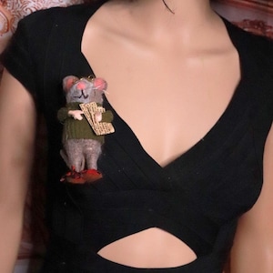Puede incluir: Un broche de ratón de lana fieltrada con cuerpo gris, orejas y nariz rosas. El ratón lleva un suéter verde, gafas pequeñas con montura dorada y sostiene un trozo de papel impreso. Se apoya sobre zapatos rojos y está prendido en una prenda negra.