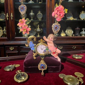 Op de afbeelding: Versierde goudkleurige ketting met een zakhorloge hanger. De ketting heeft een cherubijn, roze bloemen en blauwe accenten. Het zakhorloge toont de tijd en ligt op een fluwelen display.
