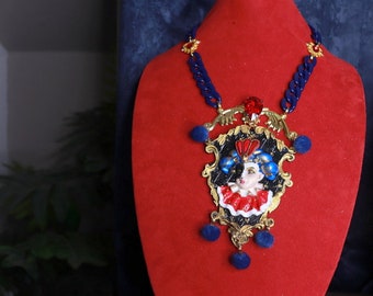 May include: A gold necklace with a blue and red pendant featuring a stylized face. The pendant is surrounded by gold filigree and has blue and red pom-poms hanging from it.