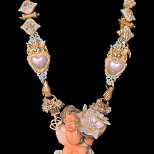 May include: A detailed necklace featuring a cherub holding a flower basket. The necklace has gold-toned accents, faux pearls, and heart-shaped pendants. The cherub is peach-colored, and the necklace has a vintage aesthetic.