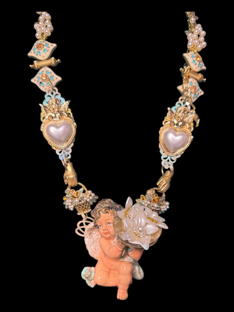 May include: A detailed necklace featuring a cherub holding a flower basket. The necklace has gold-toned accents, faux pearls, and heart-shaped pendants. The cherub is peach-colored, and the necklace has a vintage aesthetic.