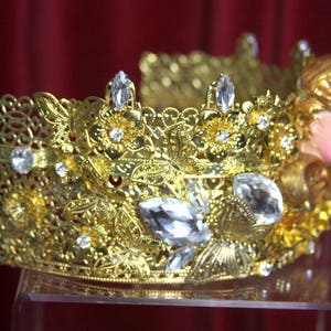 May include: A gold crown with intricate details and clear crystal accents. The crown is adorned with a small, gold cherub figure.