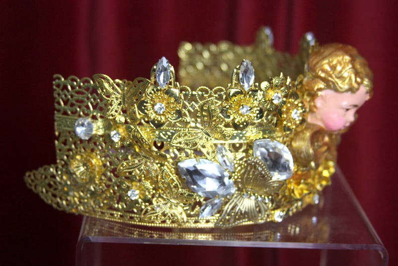 May include: A gold crown with intricate details and clear crystal accents. The crown is adorned with a small, gold cherub figure.