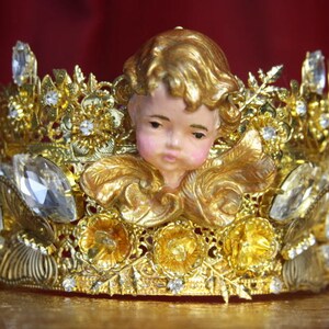 May include: A gold crown with a cherub figurine in the center. The crown is adorned with rhinestones and intricate details.