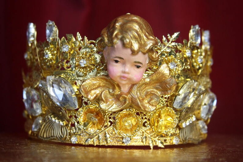 May include: A gold crown with a cherub figurine in the center. The crown is adorned with rhinestones and intricate details.