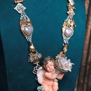 May include: A detailed necklace featuring a cherub pendant holding a decorative object. The necklace has gold-toned metal accents, faux pearls, and heart-shaped pendants. The design includes white, blue, and gold elements.