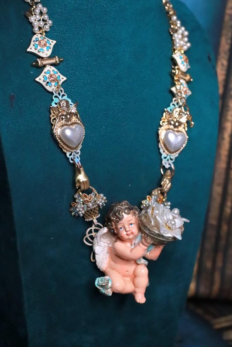 May include: A detailed necklace featuring a cherub pendant holding a decorative object. The necklace has gold-toned metal accents, faux pearls, and heart-shaped pendants. The design includes white, blue, and gold elements.