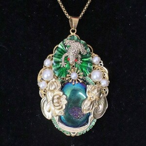 May include: A gold necklace with a pendant featuring a green frog, a iridescent blue stone, and white pearls.