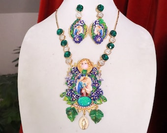 May include: A gold necklace with a large pendant featuring a religious image surrounded by green and purple flowers. The necklace is adorned with green and clear rhinestones. Matching earrings with a similar design are also included.