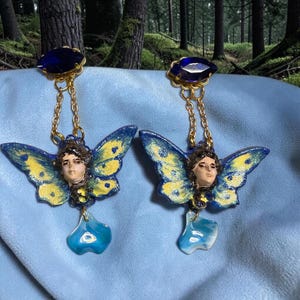 Può includere: Un paio di orecchini unici con un design a farfalla. Ogni orecchino ha una gemma blu, una catena d'oro, un viso e ali di farfalla blu e gialle. Un ciondolo blu a forma di goccia pende sotto.