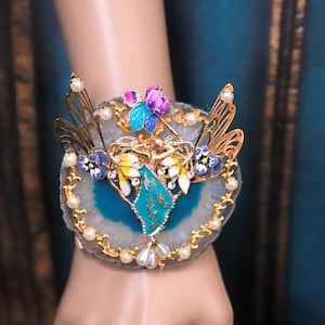 May include: A gold-tone bracelet with a blue and white agate slice, featuring a fairy figurine and butterfly wings with floral accents.