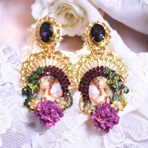 Puede incluir: Pendientes dorados ornamentados, cada uno con una gran gema ovalada púrpura. Cada pendiente presenta un retrato, rodeado de cristales burdeos, hojas verdes y una flor púrpura. Sobre un fondo de encaje blanco.