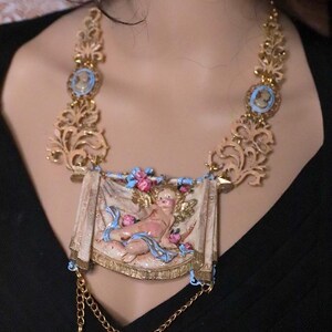 Peut inclure: Collier dor&eacute; orn&eacute; d'une banni&egrave;re centrale avec un ch&eacute;rubin, rehauss&eacute; de d&eacute;tails roses et bleus. Le collier comprend des pendentifs de style cam&eacute;e et des &eacute;l&eacute;ments de filigrane d&eacute;coratifs, cr&eacute;ant une esth&eacute;tique vintage.