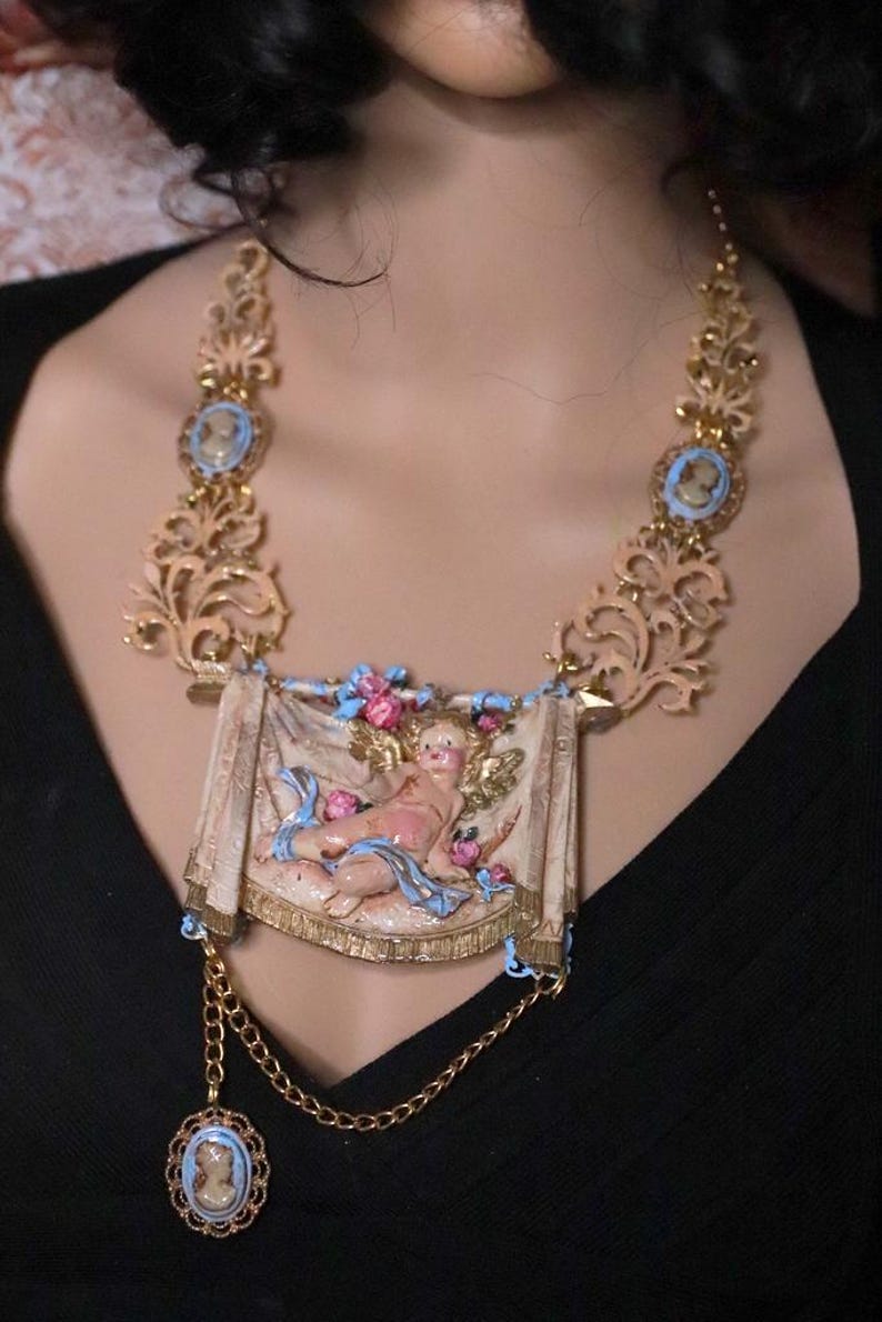 Peut inclure: Collier dor&eacute; orn&eacute; d'une banni&egrave;re centrale avec un ch&eacute;rubin, rehauss&eacute; de d&eacute;tails roses et bleus. Le collier comprend des pendentifs de style cam&eacute;e et des &eacute;l&eacute;ments de filigrane d&eacute;coratifs, cr&eacute;ant une esth&eacute;tique vintage.