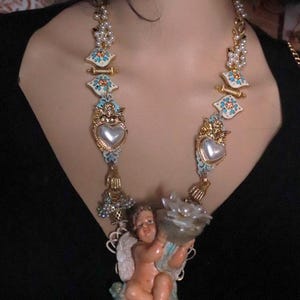 May include: A gold-toned necklace featuring ornate, decorative elements. The necklace includes heart-shaped pendants, floral accents, and a cherub figurine holding a flower-shaped object. The design incorporates pearls and turquoise-colored details.