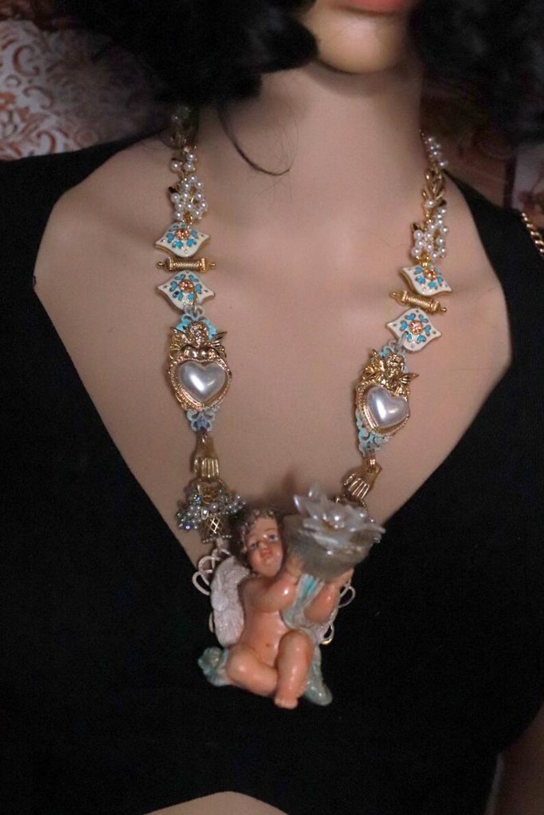 May include: A gold-toned necklace featuring ornate, decorative elements. The necklace includes heart-shaped pendants, floral accents, and a cherub figurine holding a flower-shaped object. The design incorporates pearls and turquoise-colored details.