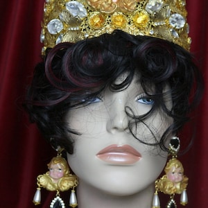 May include: A mannequin wearing a gold crown with a cherub figurine on top and large gold angel cherub earrings with black teardrop crystals.