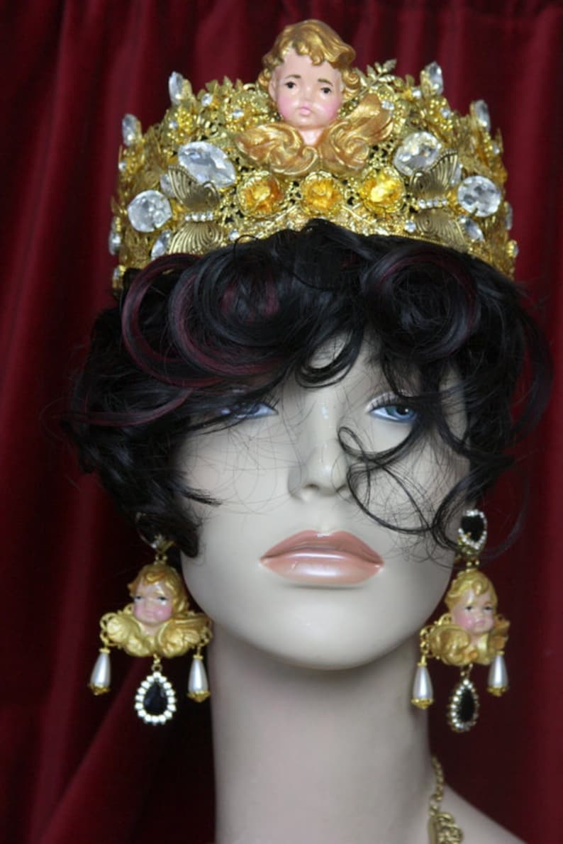 May include: A mannequin wearing a gold crown with a cherub figurine on top and large gold angel cherub earrings with black teardrop crystals.