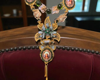 May include: Ornate gold-toned necklace with intricate floral and portrait pendant details. The necklace features a layered design with green and clear gemstone accents. The pendant showcases a portrait within a circular frame, suspended from a delicate chain.
