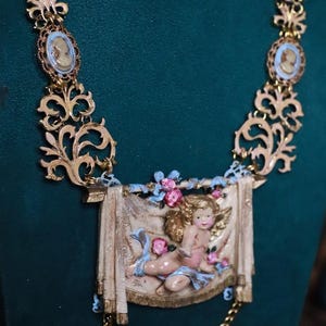 Peut inclure: Collier dor&eacute; orn&eacute; de pendentifs cam&eacute;e et d'un pendentif rectangulaire central repr&eacute;sentant un ch&eacute;rubin. Le collier pr&eacute;sente des d&eacute;tails de filigrane complexes et une cha&icirc;ne d&eacute;licate.