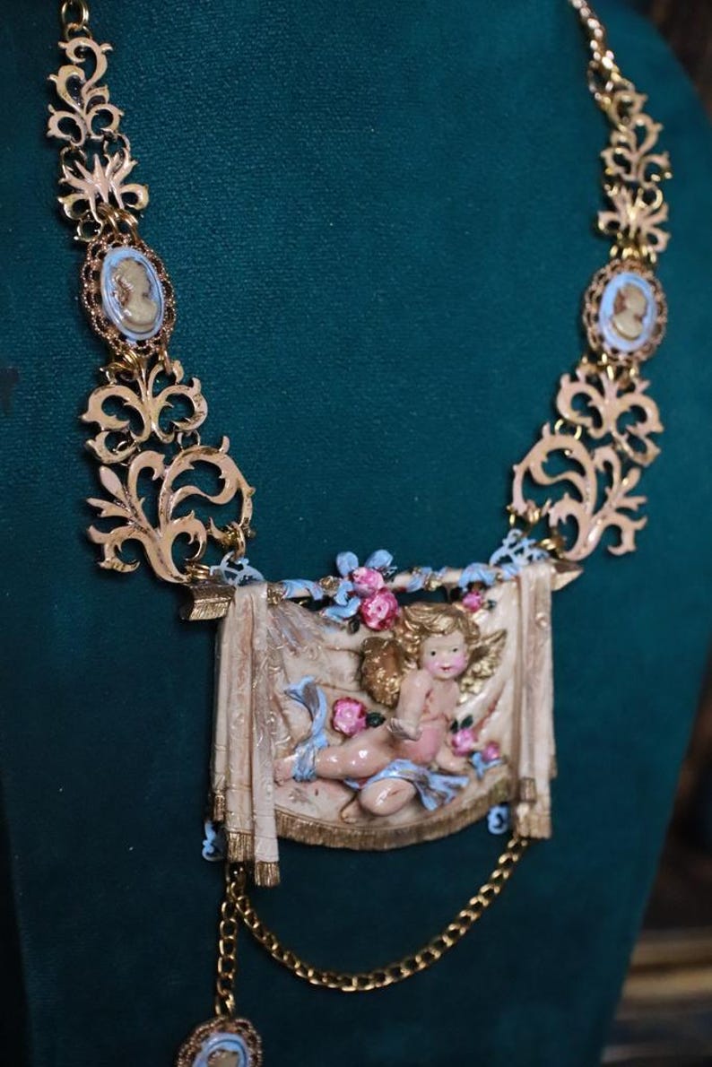 Peut inclure: Collier dor&eacute; orn&eacute; de pendentifs cam&eacute;e et d'un pendentif rectangulaire central repr&eacute;sentant un ch&eacute;rubin. Le collier pr&eacute;sente des d&eacute;tails de filigrane complexes et une cha&icirc;ne d&eacute;licate.