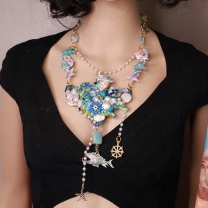 May include: A multi-layered necklace with a large, intricate pendant featuring a cluster of seashells, starfish, and a fish. The pendant is made of blue, green, and white enamel and is accented with gold-tone details. The necklace is strung on a pearl chain and includes a small gold-tone steering wheel charm.