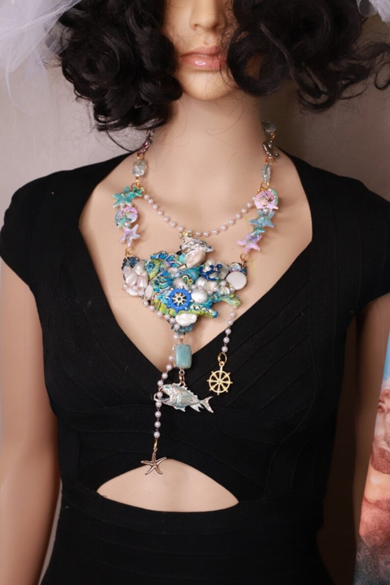 May include: A multi-layered necklace with a large, intricate pendant featuring a cluster of seashells, starfish, and a fish. The pendant is made of blue, green, and white enamel and is accented with gold-tone details. The necklace is strung on a pearl chain and includes a small gold-tone steering wheel charm.