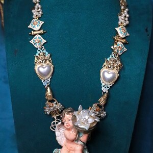 May include: Ornate gold-toned necklace featuring a cherub holding a flower-shaped object. The necklace has heart-shaped pendants, floral accents, and pearl-like beads. The design incorporates white, turquoise, and gold elements, creating a vintage aesthetic.