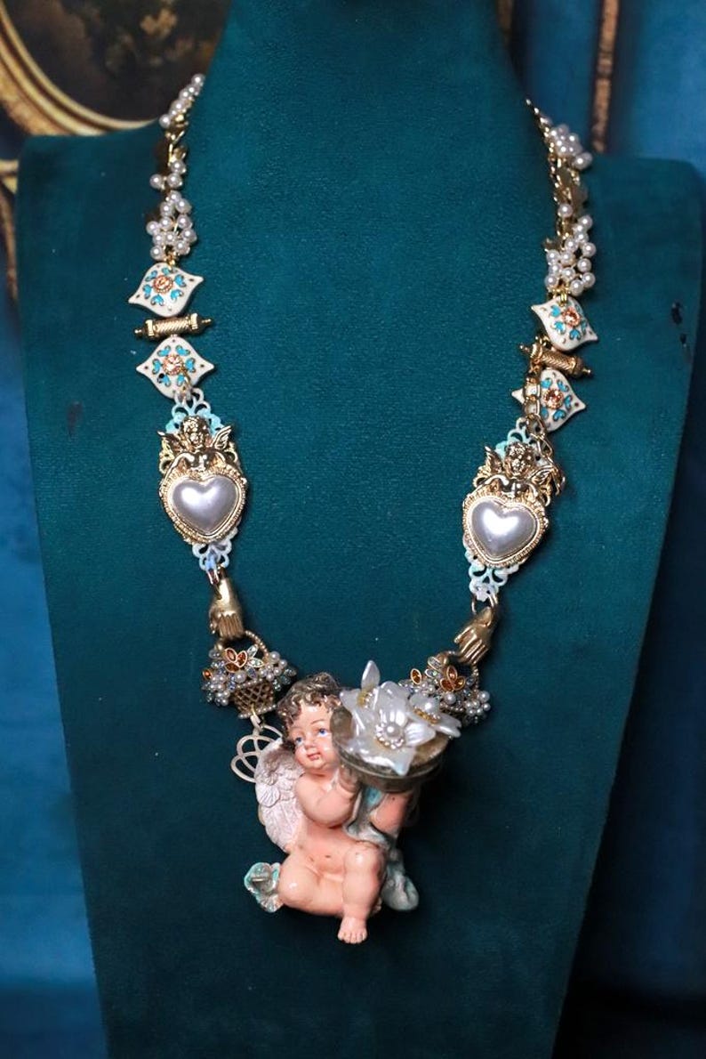 May include: Ornate gold-toned necklace featuring a cherub holding a flower-shaped object. The necklace has heart-shaped pendants, floral accents, and pearl-like beads. The design incorporates white, turquoise, and gold elements, creating a vintage aesthetic.