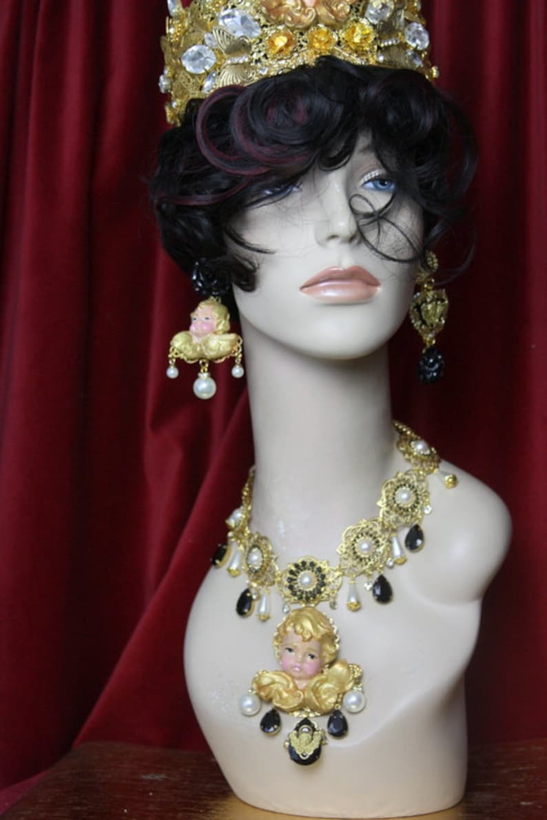 May include: A gold crown with clear and yellow rhinestones, a gold necklace with black and white beads and a cherub pendant, and matching earrings with cherub pendants.