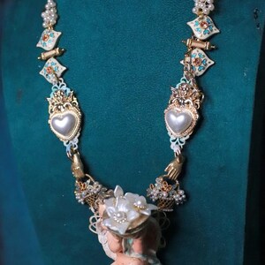 May include: An ornate necklace featuring a cherub pendant holding a floral arrangement. The necklace has heart-shaped pendants, pearl accents, and decorative gold-toned elements. The necklace is displayed against a teal background.