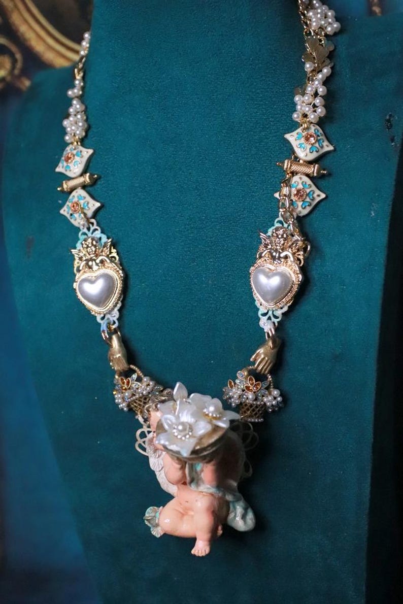 May include: An ornate necklace featuring a cherub pendant holding a floral arrangement. The necklace has heart-shaped pendants, pearl accents, and decorative gold-toned elements. The necklace is displayed against a teal background.