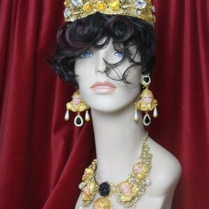 May include: A gold crown with cherub details and clear rhinestones, a gold necklace with cherub details and black accents, and matching earrings with cherub details and black accents.