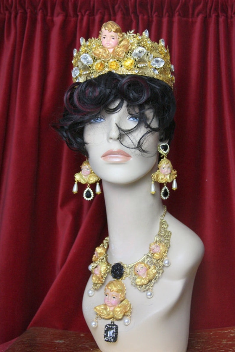 May include: A gold crown with cherub details and clear rhinestones, a gold necklace with cherub details and black accents, and matching earrings with cherub details and black accents.