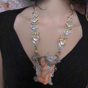 May include: A statement necklace featuring a cherub pendant holding a cornucopia. The necklace has gold-tone accents, faux pearls, and heart-shaped pendants with blue and white enamel details. The cherub is in a seated position.