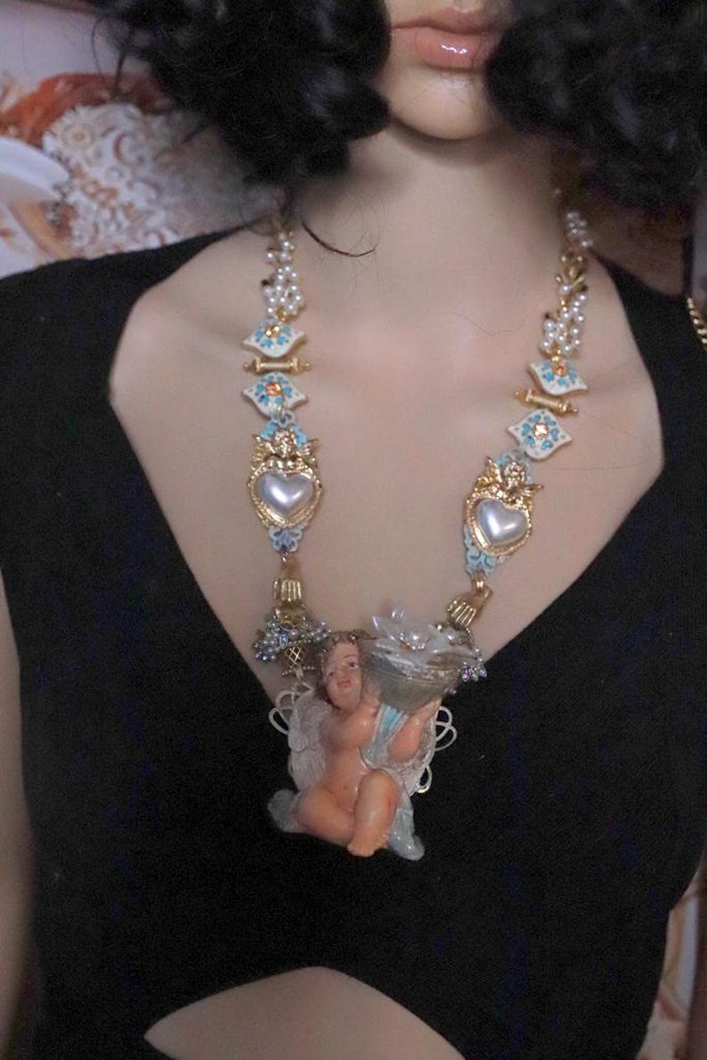 May include: A statement necklace featuring a cherub pendant holding a cornucopia. The necklace has gold-tone accents, faux pearls, and heart-shaped pendants with blue and white enamel details. The cherub is in a seated position.