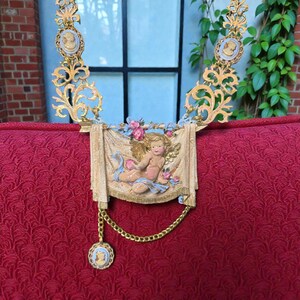Peut inclure: Collier dor&eacute; orn&eacute; de cam&eacute;es et d'un motif central de ch&eacute;rubin. Le collier pr&eacute;sente des filigranes complexes et une banni&egrave;re drap&eacute;e avec une figure de ch&eacute;rubin, orn&eacute;e de d&eacute;tails floraux roses et bleus. Une cha&icirc;ne dor&eacute;e avec un pendentif cam&eacute;e est suspendue en dessous.