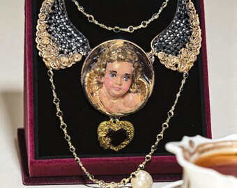 May include: A gold-toned necklace with a pendant featuring a painted cherub face. The necklace has black crystal accents and a heart-shaped charm. The necklace is displayed on a red velvet surface.