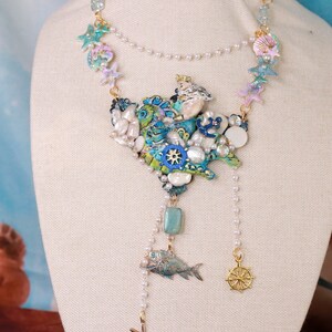 May include: A multi-layered necklace with a large, blue and green, seashell-shaped pendant. The pendant is decorated with pearls, gold accents, and a small anchor. The necklace also features a small, gold fish charm and a gold steering wheel charm.