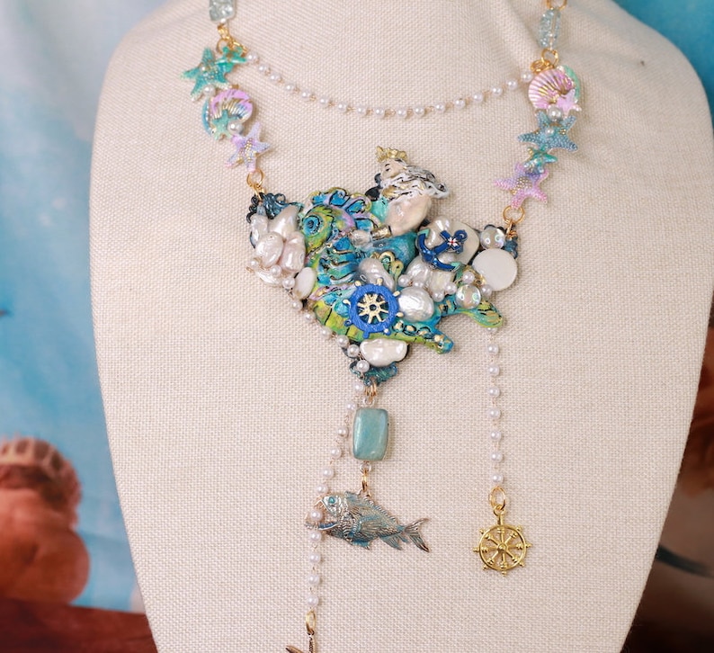 May include: A multi-layered necklace with a large, blue and green, seashell-shaped pendant. The pendant is decorated with pearls, gold accents, and a small anchor. The necklace also features a small, gold fish charm and a gold steering wheel charm.