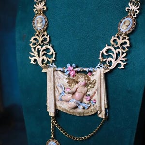 Peut inclure: Collier dor&eacute; orn&eacute; d'un pendentif central en forme de ch&eacute;rubin avec une banni&egrave;re drap&eacute;e, des fleurs et des rubans bleus. Le collier comprend des accents de cam&eacute;e et des &eacute;l&eacute;ments de filigrane d&eacute;coratifs.