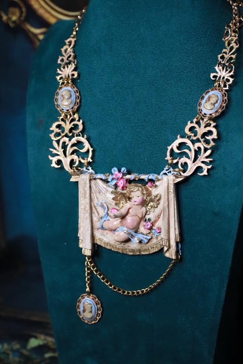 Peut inclure: Collier dor&eacute; orn&eacute; d'un pendentif central en forme de ch&eacute;rubin avec une banni&egrave;re drap&eacute;e, des fleurs et des rubans bleus. Le collier comprend des accents de cam&eacute;e et des &eacute;l&eacute;ments de filigrane d&eacute;coratifs.