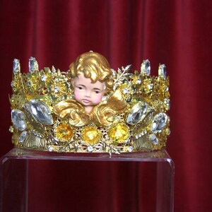 May include: A gold crown with clear rhinestones and a small porcelain cherub figurine in the center.